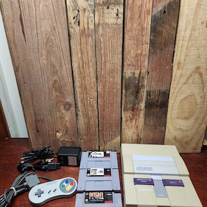 May include: A vintage Super Nintendo Entertainment System console with three game cartridges, a gray controller, and power cables. The console is beige with purple buttons. The game cartridges are gray with colorful labels.