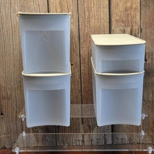 May include: Two sets of clear, square, plastic food storage containers with off-white lids. The containers are stacked on a clear acrylic display stand against a wooden background. Each container has a rectangular label area.