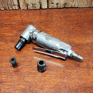 May include: A silver pneumatic right angle grinder with a black nozzle and two black collets. The tool is labeled "QUALITY TOOLS" and rests on a wooden surface.