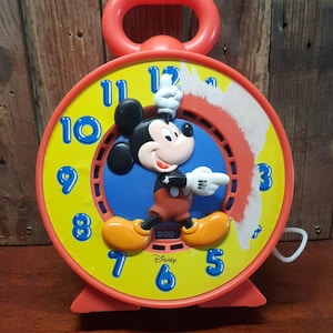 May include: A red and yellow Mickey Mouse alarm clock. The clock face is yellow with blue numbers and a red border. Mickey Mouse is in the center, with his arms raised. The clock reads 3:00 and has the Disney logo.
