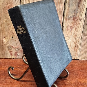 May include: A black Chain-Reference Bible with gold lettering, resting on a decorative metal stand. The book has a textured cover and a red ribbon bookmark. The stand is made of curved metal and is positioned on a wooden surface.