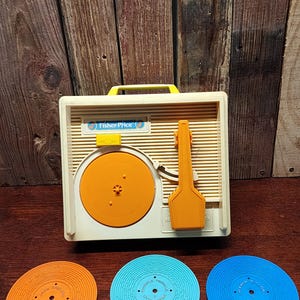 May include: Vintage Fisher-Price toy record player in off-white with an orange record and arm. Includes three colorful records: orange, light blue, and dark blue. The record player has a yellow handle and the Fisher-Price logo.