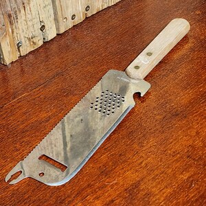 May include: A silver-colored, multi-purpose tool with a wooden handle. The tool has a serrated edge, a bottle opener, and a perforated section.