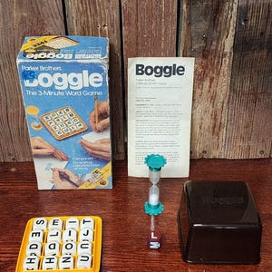 May include: A vintage Parker Brothers Boggle board game with a brown box, a green hourglass, and a black letter cube tray. The box has the game's name and logo, "Boggle: The 3-Minute Word Game". The game includes letter cubes, a timer, and a tray to hold the cubes.