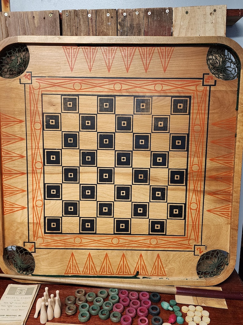 May include: A vintage wooden game board with a checkerboard pattern and orange and black accents. The board has a wooden frame and is surrounded by a rope border. The game includes wooden pieces, green and red plastic rings, and a wooden stick.