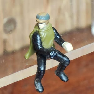 May include: A small, green and black action figure of a character wearing a brown and blue helmet. The figure is sitting on a clear surface.