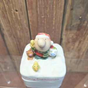 May include: A ceramic figurine featuring a character wearing a Santa hat, holding a duckling and surrounded by other small animal figurines. The base is white with the words "Tiny Treasures" in cursive.