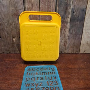 May include: A yellow plastic alphabet and number case with a handle, and a blue stencil. The case has the alphabet and numbers printed on it. The blue stencil has lowercase letters and numbers.