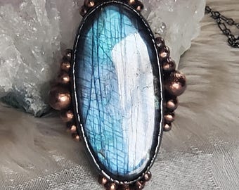 May include: Oval labradorite pendant necklace with a copper-colored metal frame and accents. The iridescent blue-green stone is set in a dark metal bezel, with a delicate chain. The pendant is approximately 2 inches long.