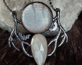 May include: A necklace featuring a circular, pale, iridescent stone set above a faceted, teardrop-shaped, off-white stone. The stones are framed by dark, textured metal branches with small bat silhouettes, suspended from a thin, dark metal chain.