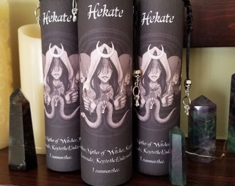May include: Three cylindrical candles with the word "Hekate" in white script. Each candle features a sepia-toned illustration of a hooded figure with keys and snakes. The candles have silver lids and decorative key charms.
