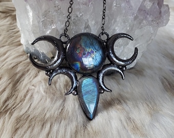May include: A dark metal pendant necklace featuring a round labradorite stone at the center, flanked by two crescent moon shapes, with a smaller teardrop-shaped labradorite stone hanging below, all suspended from a thin dark metal chain.