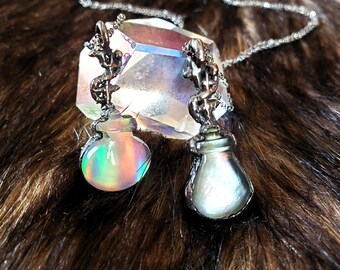 May include: Two silver-toned pendant necklaces. Each pendant features a small, iridescent glass bottle with a decorative top. The bottles are suspended from delicate silver chains. The background includes a large, faceted crystal and a dark, textured surface.
