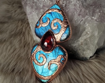 May include: A handcrafted pendant necklace featuring two blue and copper-colored pendants with a teardrop-shaped red gemstone in the center. The pendants have a swirling design and are suspended from a copper chain.