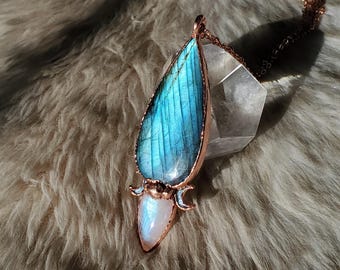 May include: A handcrafted pendant necklace featuring a large, teardrop-shaped labradorite stone with blue and green iridescence, set in a copper-colored metal frame. Below the labradorite is a smaller, pearly white moonstone. The pendant hangs from a delicate chain.
