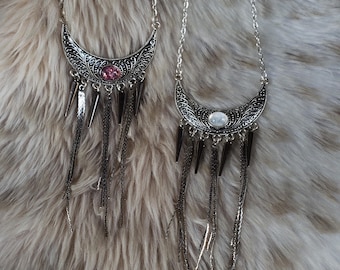 May include: Two silver-toned crescent moon necklaces featuring intricate filigree patterns. One necklace has a central pink stone, and the other has a white stone. Both pendants hang from silver chains and feature dangling metal spikes and chains.