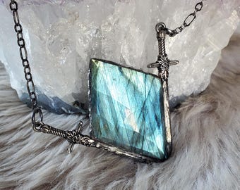 May include: A necklace featuring a faceted, diamond-shaped labradorite gemstone with iridescent blue and green tones. The stone is framed by two silver-toned metal sword motifs that connect to a dark metal chain, creating a unique pendant design.