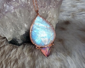 May include: A copper-colored necklace featuring a large, pear-shaped pendant with a blue and white iridescent stone. Below the pendant is a small, pink triangular stone. The necklace is displayed on a textured, light-colored surface.