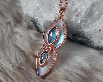 May include: A copper-colored pendant necklace featuring two teardrop-shaped sections. Each section has a central blue gemstone, surrounded by a pearly, iridescent stone. The necklace is displayed against a soft, textured background.