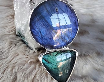 May include: A handcrafted pendant necklace featuring a large, round, blue labradorite stone above a triangular, green labradorite stone. The stones are set in a silver-colored metal frame and hang from a delicate chain.