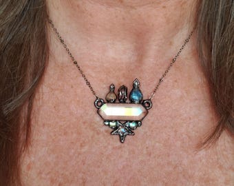 May include: A silver-toned necklace featuring a central iridescent rectangular pendant. Above the pendant are three small, decorative elements. Below the pendant is a star-shaped design with iridescent accents. The necklace is worn around the neck.