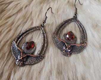 May include: A pair of drop earrings featuring a dark metallic finish with a textured, winged scarab beetle design. Each earring is centered with a faceted, deep red gemstone and hangs from a simple dark metal hook for pierced ears.