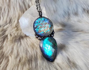 May include: A pendant necklace featuring a dark metal chain and two connected stones. The top stone is round with a carved floral pattern displaying iridescent blue and purple hues, while the bottom stone is a faceted teardrop in deep teal.