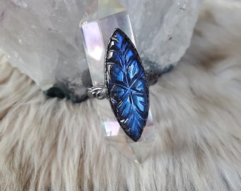 May include: A ring featuring a marquise-shaped labradorite gemstone carved with a leaf pattern. The stone displays iridescent blue flashes and is set in a dark, oxidized metal band with a twisted rope texture, resting on a clear quartz crystal.