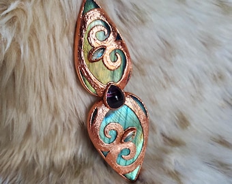 May include: A pendant necklace featuring two teardrop-shaped labradorite stones with iridescent green and blue flashes, encased in textured copper metalwork with swirling patterns and a small, dark purple gemstone set between the two sections.
