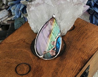 May include: A teardrop-shaped pendant with a silver-colored frame. The pendant features a colorful, iridescent stone with shades of purple, green, and blue. Delicate, rose-gold colored floral accents adorn the side. The pendant rests on a wooden box.