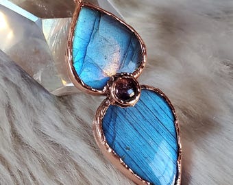 May include: A handcrafted pendant necklace featuring two teardrop-shaped, iridescent blue labradorite stones. The stones are set in a copper-colored metal frame and connected by a small, round, dark-colored stone. The necklace chain is also copper-colored.