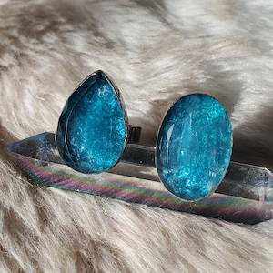 May include: A pair of silver-toned stud earrings with large, faceted, teal-colored gemstones. One earring is teardrop-shaped, the other is oval. The earrings are displayed on a clear crystal against a fluffy, white background.