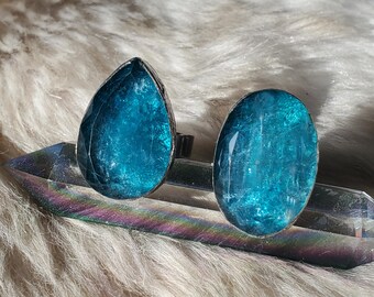May include: A pair of silver-toned stud earrings with large, faceted, teal-colored gemstones. One earring is teardrop-shaped, the other is oval. The earrings are displayed on a clear crystal against a fluffy, white background.