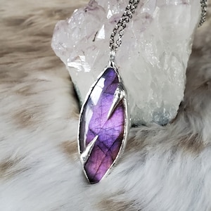 Puede incluir: Un collar de cadena de plata con un colgante de labradorita púrpura. El colgante está engastado en un marco de plata y tiene una forma única y facetada.