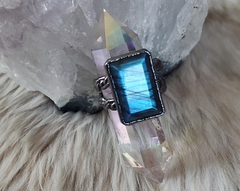 May include: A ring featuring a rectangular, faceted labradorite gemstone with a vibrant blue flash, set in a dark, oxidized metal band with a twisted rope texture. The ring is positioned around a clear, pointed quartz crystal point.