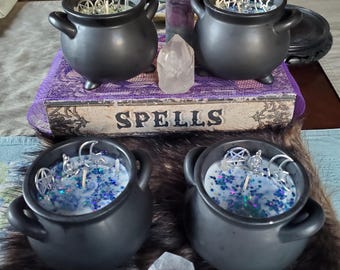 May include: Four black cauldron-shaped candles with silver charms and blue glitter. The candles sit on a purple book that reads "SPELLS". Clear quartz crystals and a purple fluorite crystal are also present.