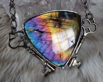 May include: A triangular labradorite pendant featuring iridescent flashes of purple, blue, and gold, set in a silver-toned metal frame with decorative key-shaped accents. The pendant is attached to a silver-toned chain with oval links.
