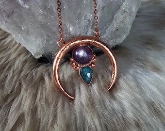 May include: A copper-toned crescent moon pendant necklace featuring a central round purple bead and a faceted teal teardrop stone. The pendant hangs from a delicate copper chain, showcasing a hammered metal texture and small decorative bead accents.