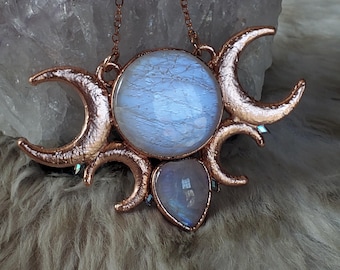 May include: A copper-toned pendant necklace featuring a large circular moonstone at the center, flanked by two crescent moon shapes on either side. A smaller, teardrop-shaped moonstone is positioned below the central stone, all set in metal.
