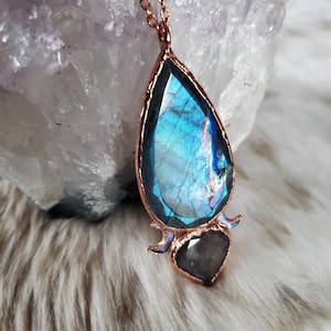 May include: A pendant necklace with a large, iridescent blue labradorite stone in a copper-coloured frame. A smaller, dark grey stone is set below the main stone. The necklace is displayed on a white, fluffy surface.