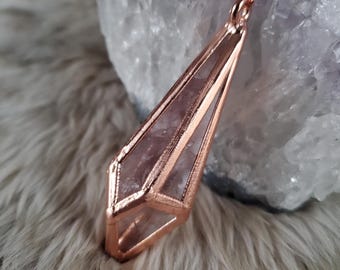 May include: A geometric pendant necklace featuring a faceted, light purple crystal encased in a copper-colored metal frame. The pendant hangs from a delicate chain, creating a modern and elegant accessory. The crystal is visible through the open design.