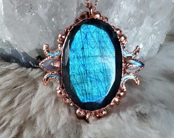 May include: A rose gold pendant necklace featuring an oval labradorite gemstone with blue iridescence. The pendant is framed by a decorative rose gold setting with small, round accents and blue-toned embellishments. The necklace is displayed on a soft, textured surface.