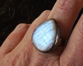 May include: A pear-shaped moonstone ring with a silver band. The moonstone has a blue and white iridescent glow. The ring is worn on a finger.