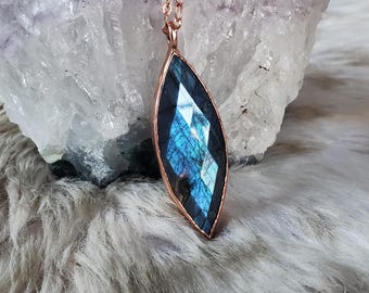 May include: A marquise-shaped labradorite pendant featuring a faceted surface with iridescent blue flashes, encased in a copper metal frame. The pendant hangs from a delicate copper chain, showcasing a polished finish and geometric design.