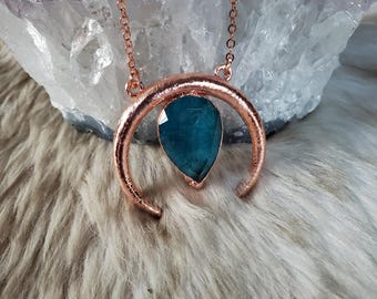 May include: A necklace featuring a rose gold-toned crescent moon pendant with a textured surface. A single, faceted, pear-shaped teal gemstone is suspended from the center of the crescent. The pendant hangs from a delicate rose gold-toned chain.
