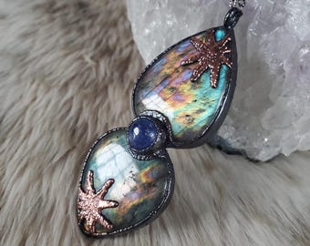 May include: A handcrafted pendant necklace featuring two iridescent labradorite teardrop-shaped stones connected by a small blue stone. The pendant has copper star accents and a dark chain. The necklace is displayed on a fluffy, light-colored surface.