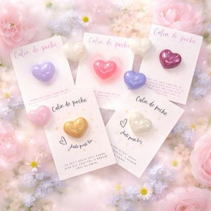 Könnte beinhalten: Eine Sammlung herzförmiger Taschenumarmungen in verschiedenen Farben, darunter Lila, Rosa, Gold und Weiß, auf Karten mit französischem Text. Der Hintergrund ist floral und weich.