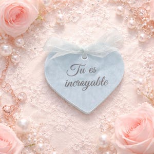 Puede incluir: Una etiqueta en forma de corazón azul claro con el texto "Tu es incroyable", atada con una cinta azul claro. La etiqueta está rodeada de rosas rosas, perlas y un delicado fondo de encaje.