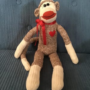 May include: A handmade sock monkey doll with brown and white striped body, white limbs, and a red mouth. It has a red heart on its chest and a red ribbon around its neck. The monkey is sitting on a blue surface.