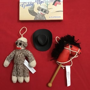 May include: A collection of cowboy-themed toys and a flipbook on a red surface. The toys include a sock monkey, a black cowboy hat, and a red stick horse with black mane. The flipbook is titled "Giddy Up!"